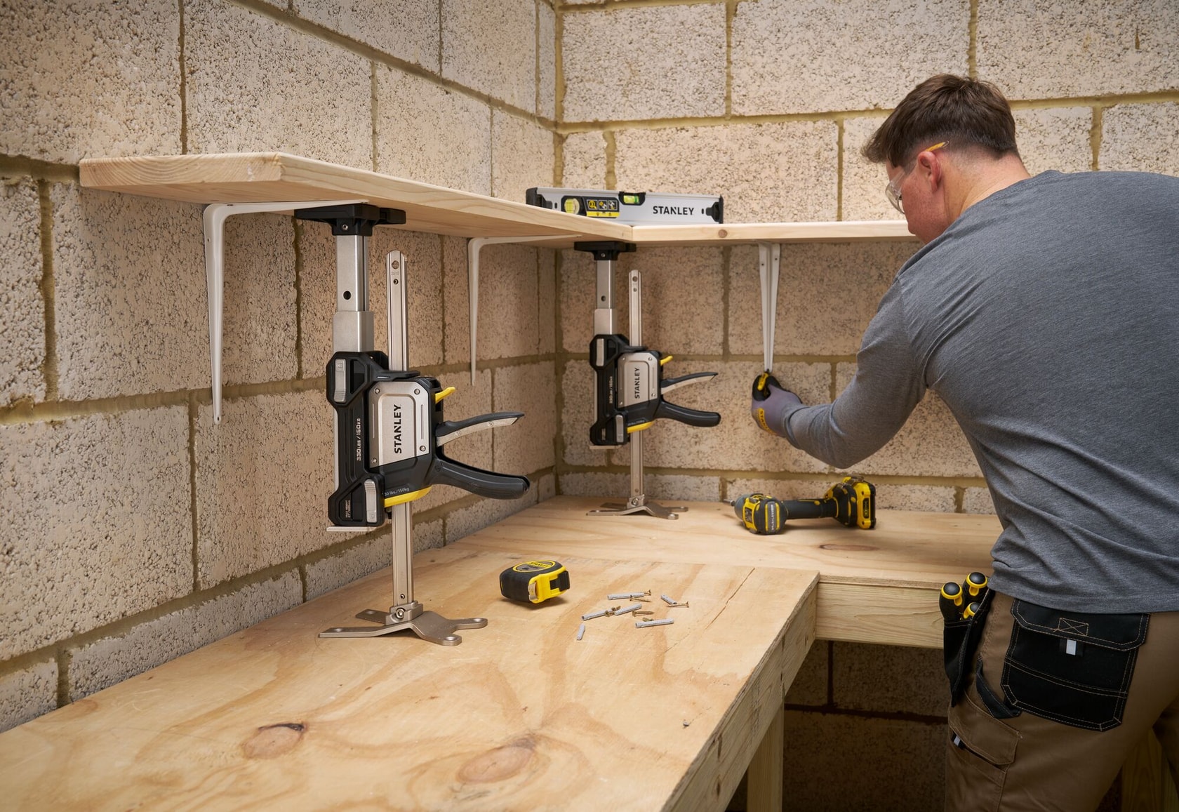STANLEY FMHT83554-1 serre-joint fixant une étagère en bois, atelier avec outils et homme assemblant, vue de face.